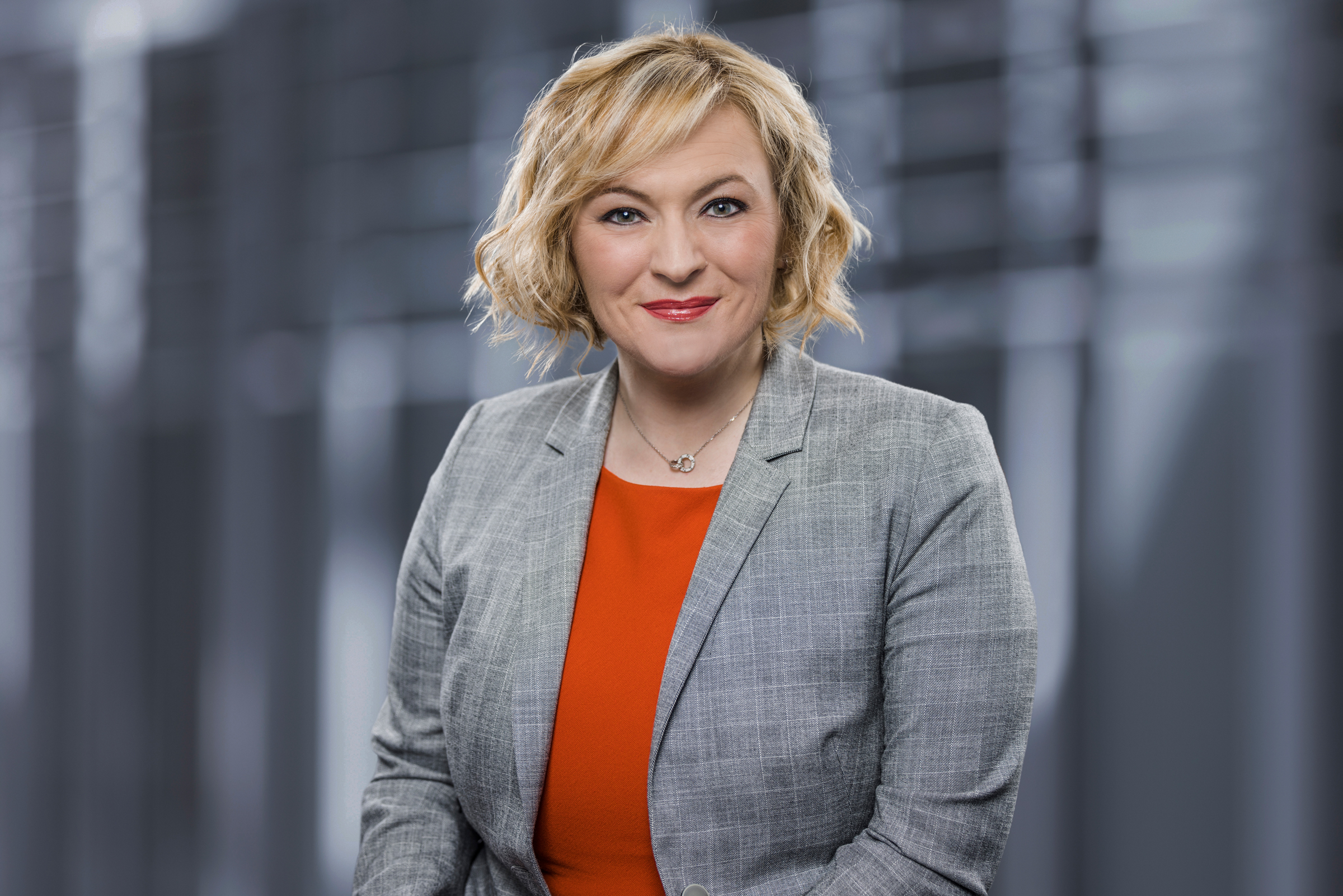 Frau mit kurzem blondem Haar in grauem Blazer und rotem Top steht als Rechtsanwältin vor modernem Hintergrund.